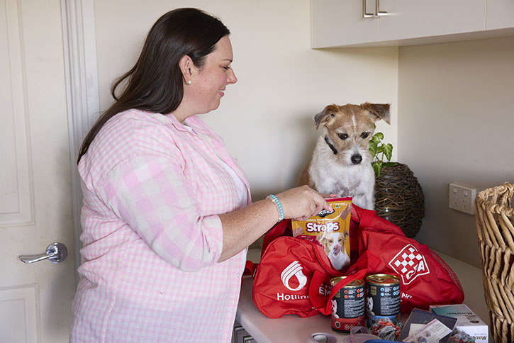 A woman is packing a bag with dog food, treats and medication. Her dog is with her and she is smiling.